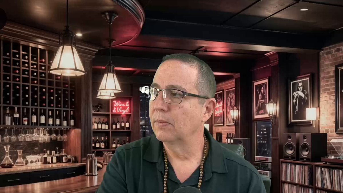 In the image, there is a man sitting at a bar with glasses on his face. He appears to be looking off into the distance or thinking about something. The bar has a wine rack filled with bottles behind him, indicating that he might have been enjoying some drinks earlier. There are also several books visible in the scene, suggesting that this could be a cozy and intellectual atmosphere for patrons.