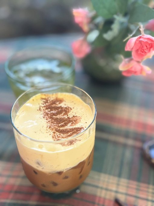 A close-up photo of a layered iced coffee drink with a creamy foam topping and a sprinkle of cinnamon, with pink flowers in the background.