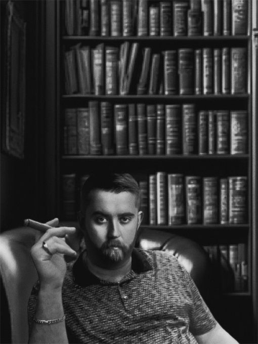 Black and white portrait of a bearded man wearing a textured polo shirt, holding a cigar while seated in front of a bookshelf filled with books.