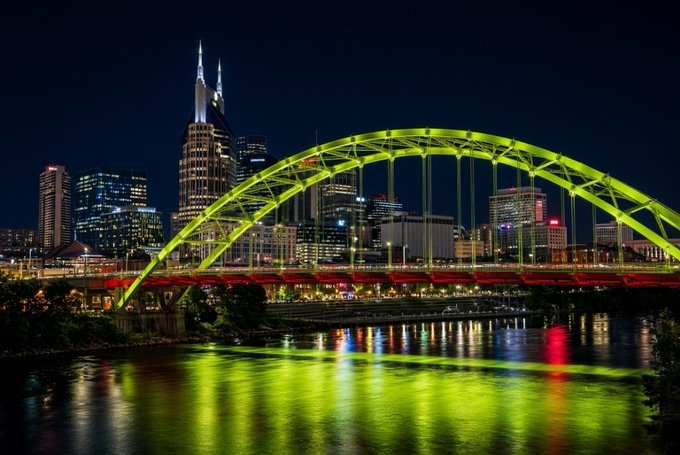 A nighttime cityscape featuring a brightly lit yellow and red arch bridge over a river, with a city skyline in the background.