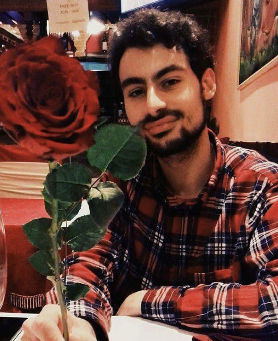 A young man with a beard, wearing a red plaid shirt, holds up a large red rose while smiling softly at the camera in what appears to be a restaurant setting. The caption "Me all lovey dovey in 2017 instead of buying bitcoin — Fml" humorously frames the romantic photo as a missed financial opportunity during bitcoin's early rise.