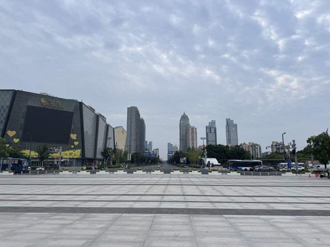 A cityscape view of a large plaza with modern skyscrapers, a building with a digital screen and yellow heart decorations, and a street leading into the urban skyline under a partly cloudy sky.