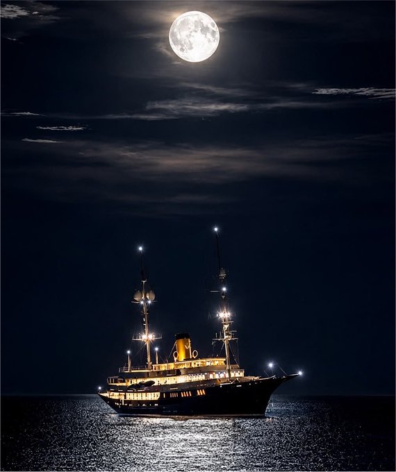 A scenic nighttime photograph of a large illuminated ship sailing on calm waters beneath a bright full moon.