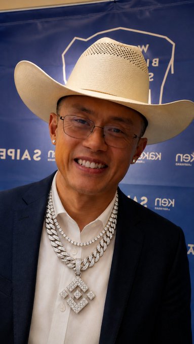 A man wearing a white cowboy hat, glasses, a dark suit, and a large diamond-encrusted pendant necklace, smiling in front of a blue background with logos.