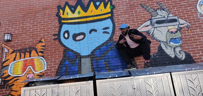 A man crouches in front of a colorful street mural featuring a crowned blue cat-like character, an orange tiger with goggles, and a unicorn with a tongue out.