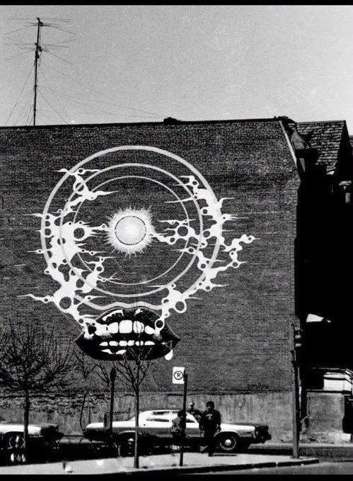 A black and white photograph of a brick building wall featuring large surreal graffiti depicting a stylized eye within a circle above lips with teeth, with vintage cars and people on the street below.