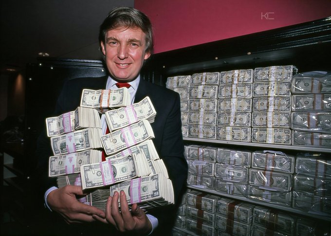 A man in a suit holds several thick stacks of US hundred-dollar bills in front of a shelf overflowing with even more money. The image serves as a visual representation of wealth intended to reinforce the accompanying post's message about becoming a millionaire.