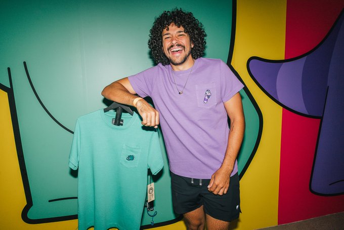 A smiling man with curly hair wearing a purple t-shirt, standing in front of a colorful wall mural while holding up a teal t-shirt on a hanger.