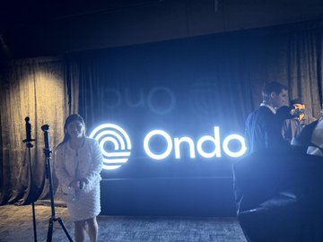 Two people stand near a glowing 'Ondo' sign on a stage with dark curtains in the background.