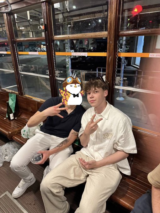 Two young men sitting on a wooden bench inside what appears to be a tram, one with a penguin sticker over his face, both making peace signs.