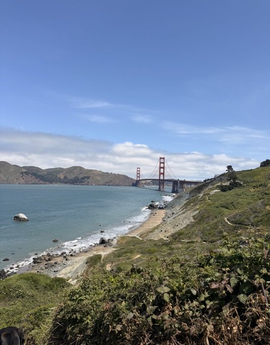 A scenic coastal view of the Golden Gate Bridge in San Francisco, with sandy beaches, blue water, and hills in the foreground under a partly cloudy sky.
