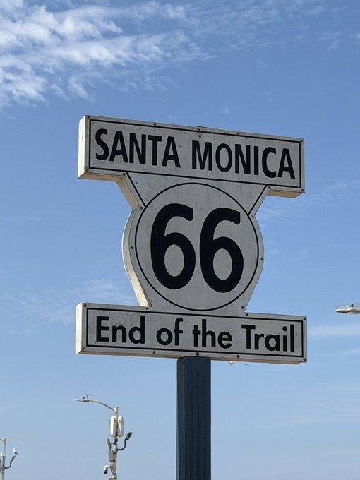 The iconic 'SANTA MONICA 66 End of the Trail' sign against a partly cloudy blue sky, marking the western terminus of the historic Route 66.