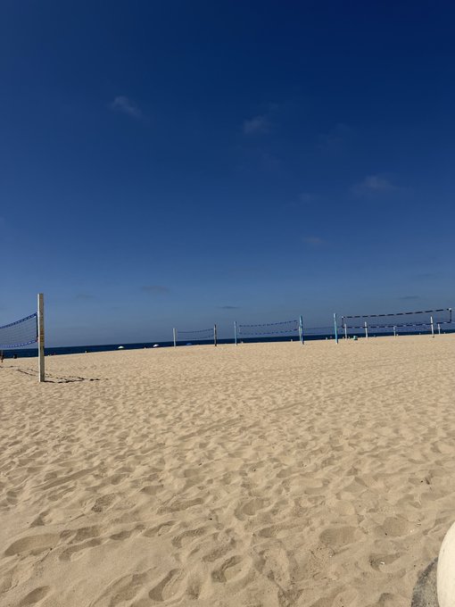 A serene beach scene features volleyball nets set against an expansive sandy shore under a deep blue, mostly cloudless sky. The calm, untouched sand and clear horizon evoke the peaceful, beautiful day promised in the morning greeting.