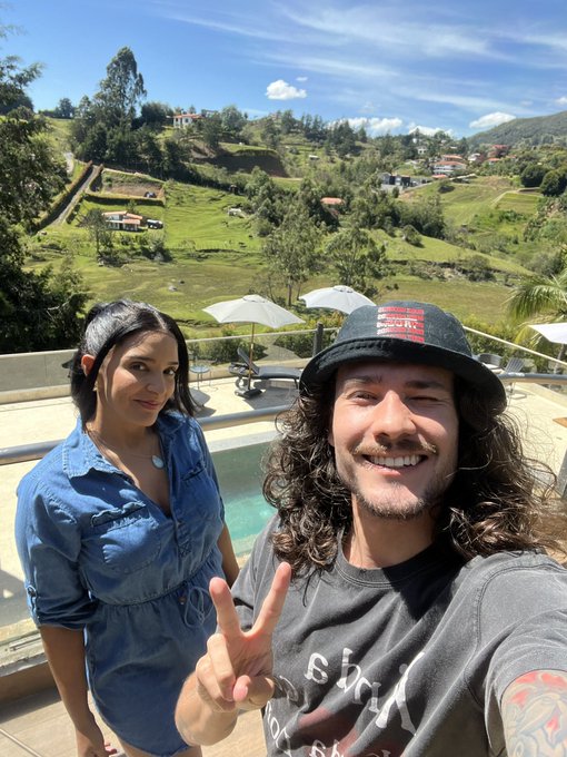 A smiling couple taking a photo by a pool, with the man wearing a bucket hat and dark t-shirt flashing a peace sign, and the woman in a blue denim shirt, set against a backdrop of green hills and houses.