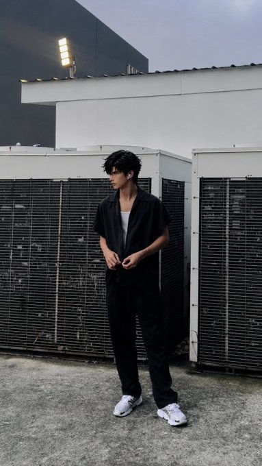 A young man in a black short-sleeved shirt and matching trousers stands on a rooftop near large industrial AC units, looking off to the side.