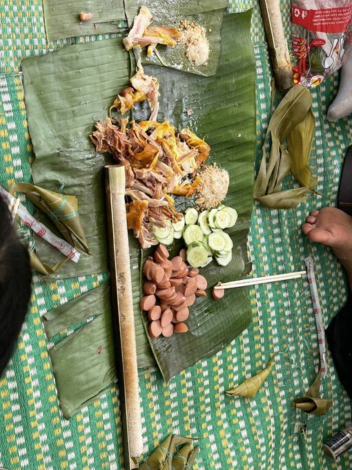 A traditional Southeast Asian meal is spread across banana leaves on a woven mat, featuring shredded chicken, sliced cucumbers, sausages, rice, and various wrapped parcels, with an "OREO" package visible at the top. The communal, shared nature of this feast mirrors the post's message about seamless, borderless payments that KAST is building with stablecoins—where diverse elements come together effortlessly, just like the different dishes gathered on one leaf.