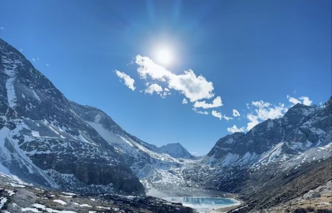 A bright sun shines over a snowy mountain valley with a small blue-green lake nestled between rugged peaks.