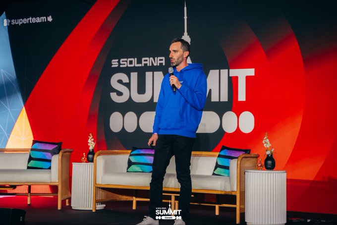 A man in a blue hoodie speaks into a microphone on stage at the Solana Summit, with 'SOLANA SUMMIT' visible on the backdrop.