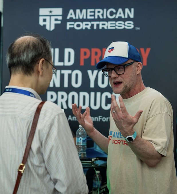A man in a blue and white 'American Fortress' cap and tan t-shirt gestures with both hands while speaking to another man at a booth with an 'AMERICAN FORTRESS' banner reading 'BUILD PRIVACY INTO YOUR PRODUCTS'.