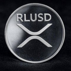 A silver commemorative coin featuring the XRP logo with 'RLUSD' engraved at the top, representing the Ripple USD stablecoin.