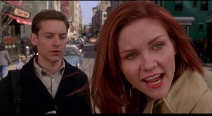A movie still featuring Tobey Maguire and Kirsten Dunst on a busy city street, with Kirsten in the foreground appearing to speak while Tobey stands behind her.