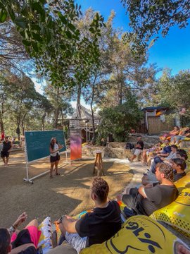 An outdoor gathering in a wooded area where people sit on a hillside watching a presentation, with a person standing near a chalkboard and a small dog visible in the center.