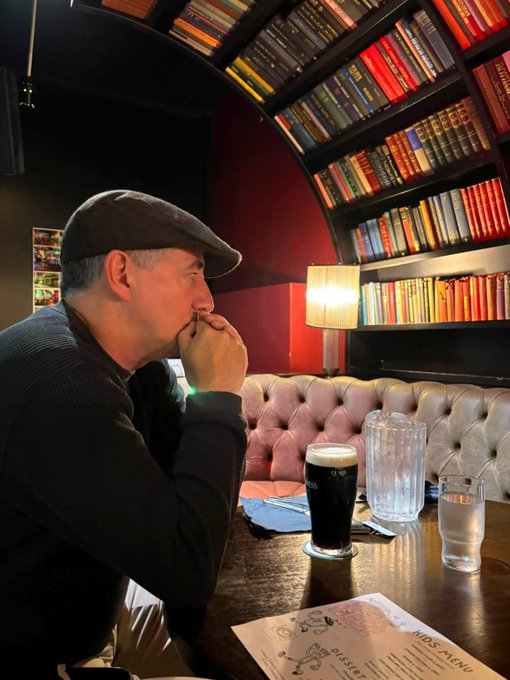 A man in a flat cap sits thoughtfully at a pub table with a dark beer, a water pitcher, and a dessert menu visible, while his mind is elsewhere—debating the investment split between $ASST and @Strategy, despite being physically in Ireland with his wife.