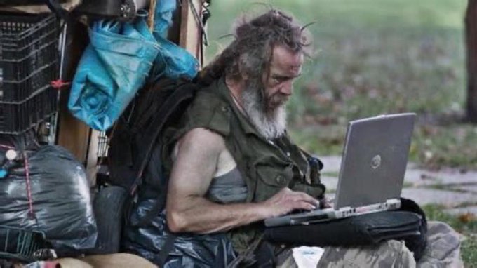 A scruffy, bearded man with long hair, wearing layered makeshift clothing and gloves, sits outdoors beside trash bags and crates, intently typing on a Dell laptop. The image humorously contrasts his rugged, "off-grid" appearance with the relatable caption "we are so fucking back," implying that despite looking like a wilderness hermit, he's connected to internet culture just like everyone else.