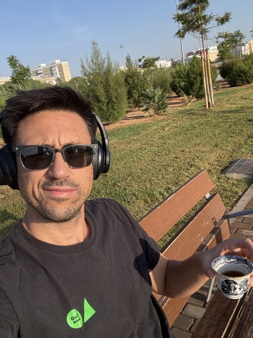 A man wearing sunglasses, headphones, and a black t-shirt sitting on a wooden bench outdoors, holding a small decorative teacup.