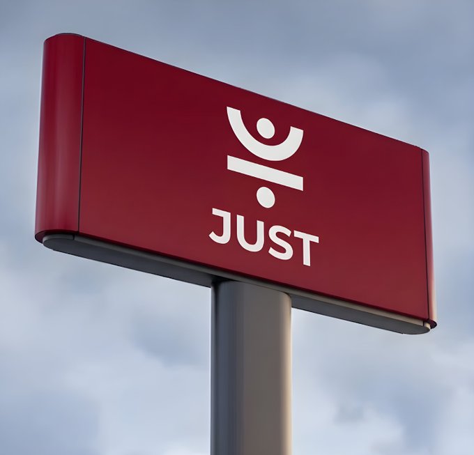 A red sign or billboard mounted on a pole against a cloudy sky, displaying a white stylized logo and the word 'JUST'.