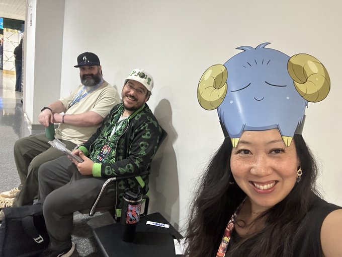 A woman in the foreground wears a cartoon blue ram mask with yellow curled horns, smiling, while two men sit behind her, one wearing a green lantern-themed outfit and cap, and another in a black cap and tan shirt at what appears to be a convention.