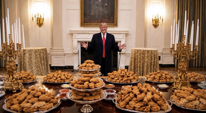 A photo of Donald Trump standing with arms outstretched in an ornate room with white columns and gold candelabras, behind a long table covered with numerous silver platters piled high with fried chicken.