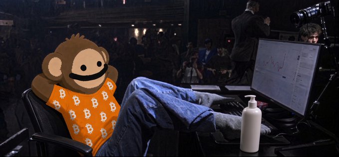 A person wearing a Bored Ape NFT avatar t-shirt and jeans sits casually with their feet up on a desk while looking at a stock chart on a computer monitor.