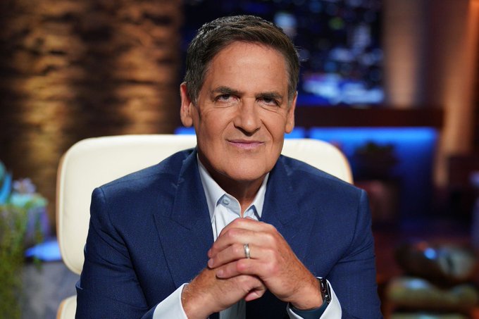 A portrait of entrepreneur Mark Cuban wearing a blue suit jacket and white shirt, sitting in a white chair with his hands clasped, smiling at the camera in what appears to be a TV studio setting.