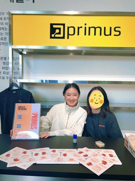 A photo of two smiling women standing behind a counter at a 'primus' store display, with various small product packages spread out on the counter in front of them.