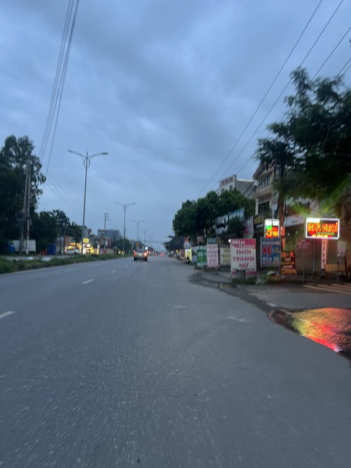 A dusk-time street view of a road lined with streetlights, trees, and various shop signs with Vietnamese text, including a sign reading 'THỜI TRANG NỮ', under a cloudy sky.