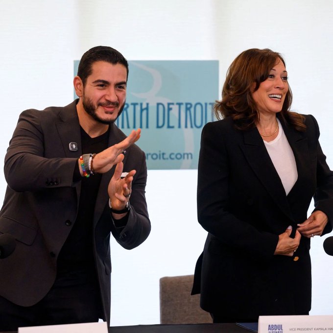 Abdul El-Sayed, identified by a nameplate as Vice President of Kamala Harris's campaign, gestures enthusiastically while standing alongside Kamala Harris, who laughs warmly during a North Detroit event featuring signage for "North Detroit" and "detroit.com."
