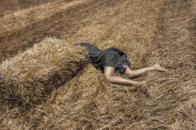 An artistic photograph showing a person lying face down in golden hay inside a barn, with a piece of dark fabric partially covering their upper body.