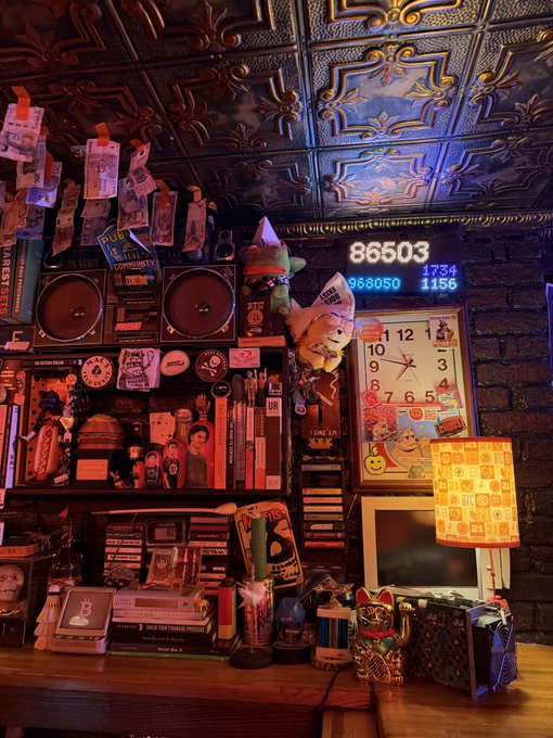 A dimly lit, cluttered shelf filled with books, stickers, a clock, small figurines, and a digital display showing '86503' and other numbers under warm red lighting.