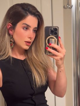 A woman with long blonde hair and statement earrings takes a mirror selfie while wearing a black sleeveless top, holding a phone with a clear case and red manicured nails. The "good morning" caption sets a cheerful tone to begin the day, inviting followers to share their own art or artistic expressions.