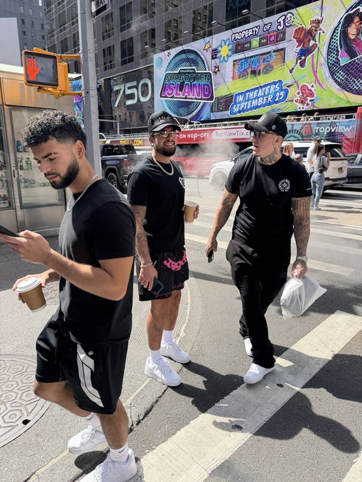 Three young men in black clothing cross a New York City street, with one wearing a shirt reading 'Property of... 16' and another with 'Arc' logo; a billboard advertises 'Forgotten Island' in theaters September 15.