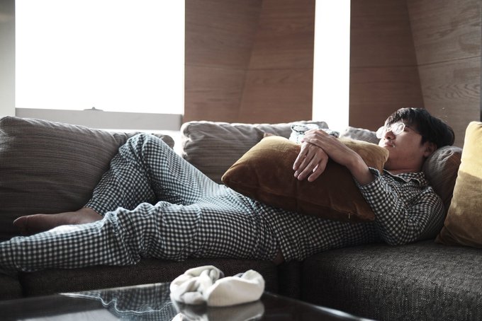 A man in checkered pajamas lounges on a couch, resting against a brown pillow in a relaxed pose. The image contrasts with the accompanying critical post, depicting an innocent, casual moment that the commentary suggests masks a calculated "sweet" persona.