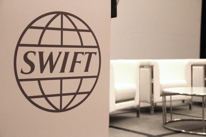 The SWIFT logo is prominently displayed on a wall in what appears to be a modern lounge or office setting, with blurred seating and a coffee table visible in the background. The image accompanies news of SWIFT announcing it will test blockchain technology on its $1.5 quadrillion messaging network, visually representing the financial messaging giant at the center of this development.