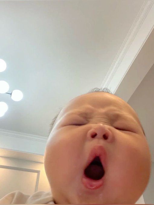 A close-up photo of a baby with eyes closed and mouth wide open, appearing to yawn, taken from a low angle with a modern ceiling light fixture visible in the background.