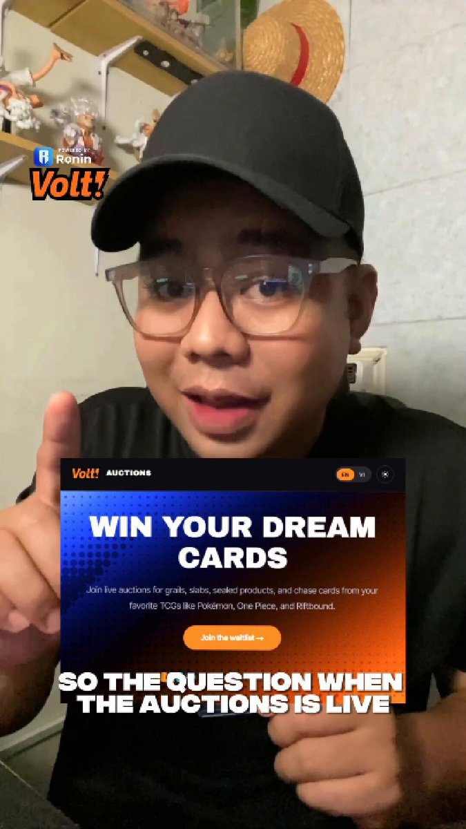 The image features a man wearing glasses holding up an electronic device with a message about winning a Pokémon card game. The man is dressed in a black shirt and hat, and he appears to be posing for a photo or video. The background of the image shows a wall with shelves containing various items such as figurines and toys.