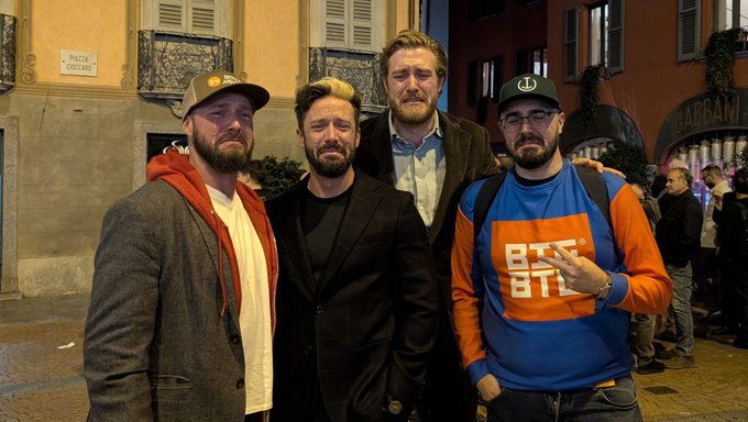 Four men pose on a city street at night, with a wall plaque reading “PIAZZA TOSCANI” visible behind them.