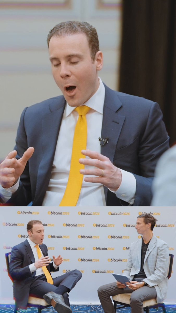 A man in a blue suit and yellow tie speaks at a conference, accompanied by another seated individual, with a backdrop of a white wall adorned with logos for "Bethany" and "Dotcom".