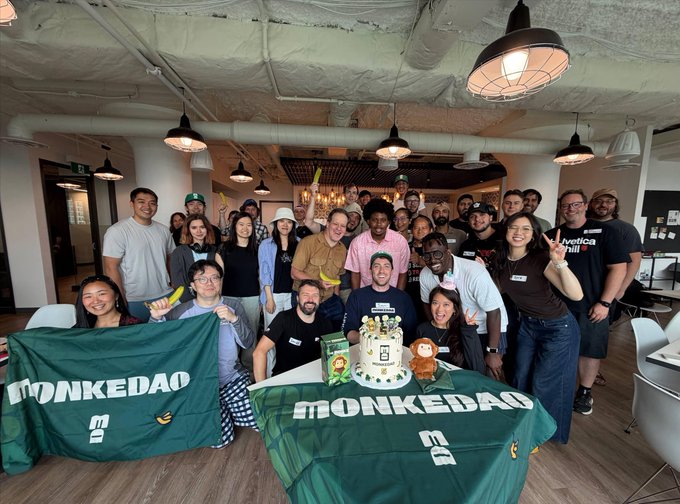 A group photo of people gathered around a table with a 'MONKEDAO' branded cake, holding green MONKEDAO flags in what appears to be an office or coworking space.