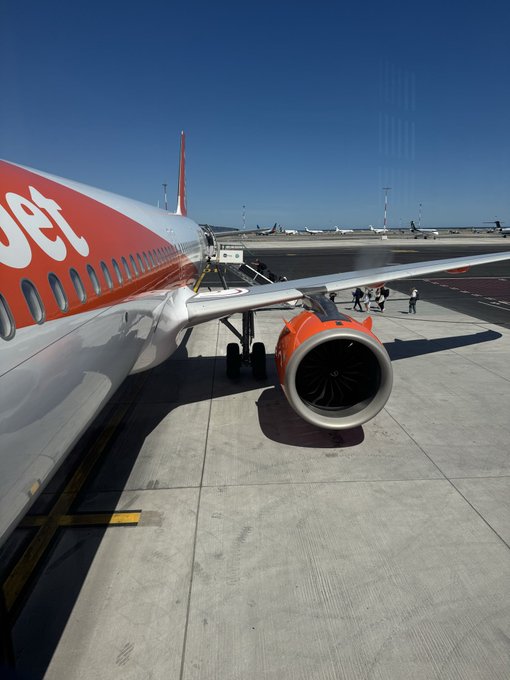 The image shows a large airplane parked at an airport, with its landing gear down. There are several people walking around near the plane, possibly preparing for their flight or just arriving from one. The scene captures the essence of air travel as passengers go about their journeys.