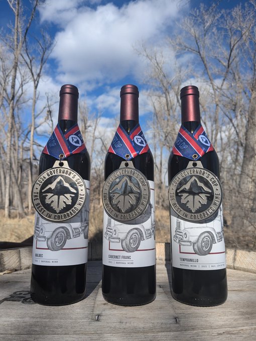 Three bottles of 'The 2024 Governor's Cup Best in Colorado Wine' arranged on a wooden surface outdoors. The bottles are labeled 'Malbec', 'Cabernet Franc', and 'Tempranillo', each featuring a silver medal-style tag around the neck.